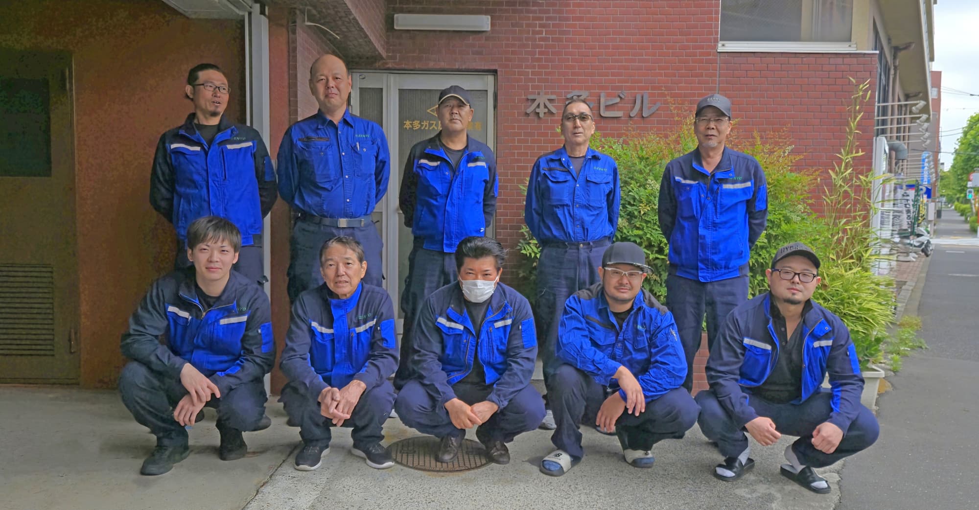 会社の前で笑顔でポーズをとる青い作業着を着た10人の男性従業員